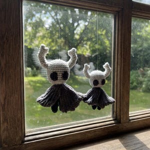 Könnte beinhalten: Zwei handgefertigte Häkelfiguren inspiriert von der Hollow Knight-Figur, mit hellgrauen, abgerundeten Köpfen, dunkelgrauen ovalen Augen und markanten Hörnern, befestigt an dunkelgrauen Umhängen. Die Figuren hängen an dünnen, transparenten Fäden.