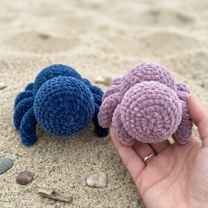 May include: Two plush, crocheted spiders in blue and lavender sit on sand. The blue spider is on the left, and the lavender spider is held in a hand on the right. The spiders have round bodies and eight legs.