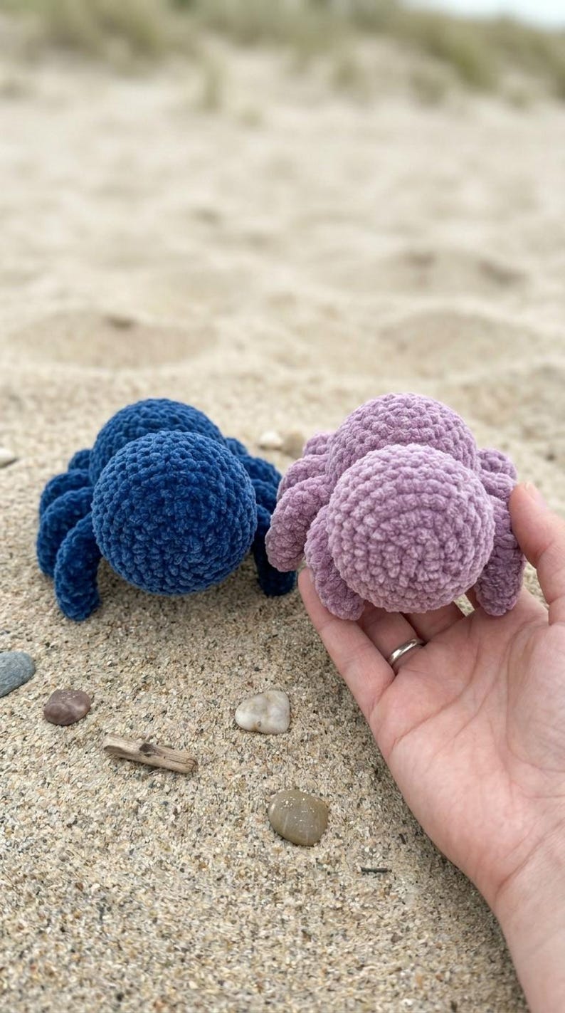 May include: Two plush, crocheted spiders in blue and lavender sit on sand. The blue spider is on the left, and the lavender spider is held in a hand on the right. The spiders have round bodies and eight legs.