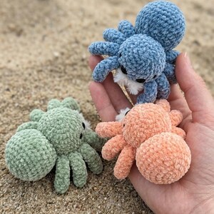 May include: Three crocheted octopus toys in shades of blue, peach, and green. The plush toys have eight legs and small black eyes. The octopus toys are displayed on a sandy surface.