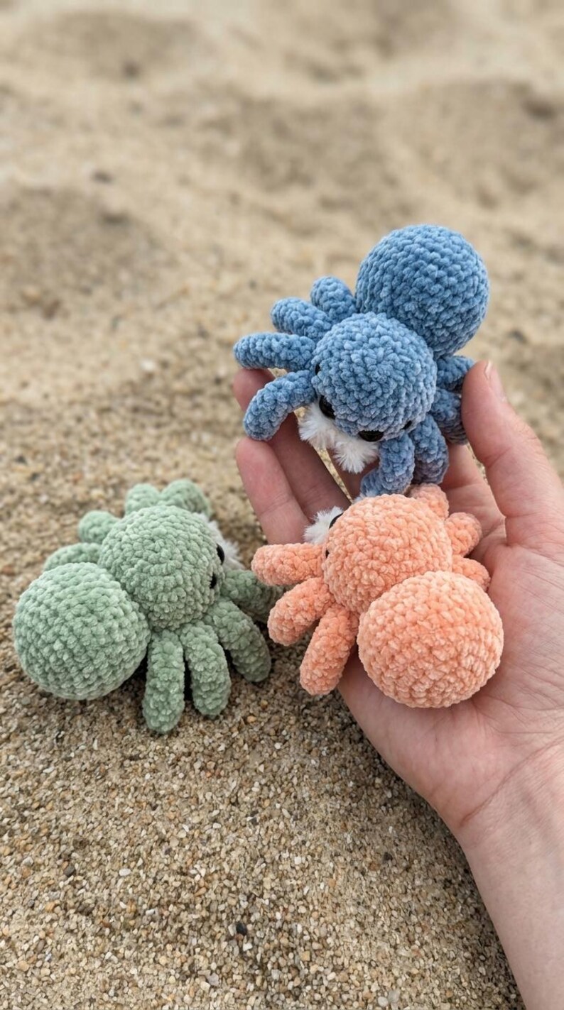 May include: Three crocheted octopus toys in shades of blue, peach, and green. The plush toys have eight legs and small black eyes. The octopus toys are displayed on a sandy surface.