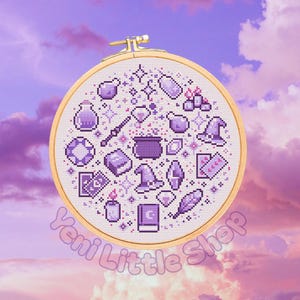 May include: A circular wooden embroidery hoop containing a white fabric piece with purple cross-stitched icons including a cauldron, potion bottles, books, crystals, a wand, a hat, and playing cards. The text Yeni Little Shop is visible below.