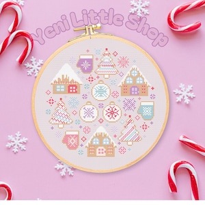 May include: A cross-stitch embroidery hoop featuring a festive winter design with houses, trees, ornaments, and mittens in pastel colors. The text "Yeni Little Shop" is arched above the hoop. Candy canes and snowflakes surround the hoop on a pink background.
