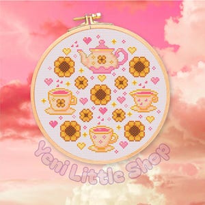 May include: Cross-stitch embroidery in a wooden hoop features a pink teapot, teacups, and sunflowers. The design includes pink hearts and yellow accents on a white background. The text "Yeni Little Shop" is at the bottom.
