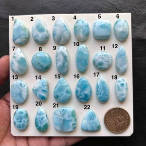 Op de afbeelding: Een verzameling van tweeëntwintig gepolijste blauwe Larimar-cabochons gerangschikt op een wit displaybord, genummerd van één tot tweeëntwintig, met een dollar-munt voor schaal om de grootte van de stenen te tonen.