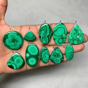 Könnte beinhalten: Eine Sammlung von zehn polierten Malachit-Edelsteinen in verschiedenen Formen, darunter rund, dreieckig und oval. Die Edelsteine zeigen eine leuchtend grüne Farbe mit einzigartigen Wirbelmustern. Die Malachitsteine werden in einer Hand gehalten.