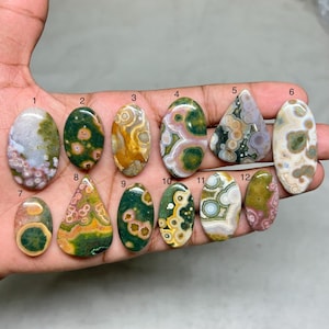 Könnte beinhalten: Eine Sammlung von zwölf polierten Edelsteinen in verschiedenen Formen, die in einer Hand gehalten werden. Die Steine zeigen eine Reihe von Farben, darunter Grün, Braun, Weiß und Rosa, mit einzigartigen Mustern und Texturen. Jeder Stein ist nummeriert.