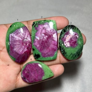 May include: Four polished ruby in zoisite gemstones. The gemstones have a vibrant pinkish-red centre surrounded by a green exterior. The shapes include oval, rectangular, and teardrop. The gemstones are displayed on a neutral background.