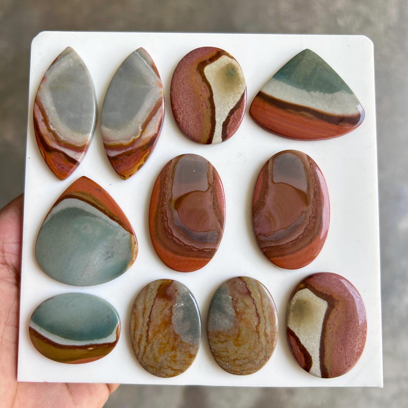May include: A collection of polished gemstones in various shapes, including ovals, teardrops, and triangles. The stones display a range of colors, such as red, brown, gray, and cream, with unique swirling patterns. The gemstones are arranged on a white surface.