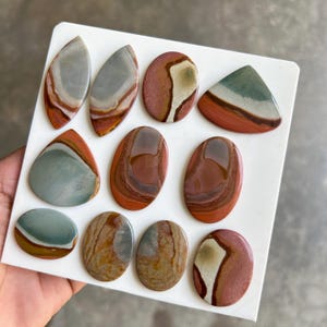 May include: Assortment of polished jasper stones in various shapes, including oval, teardrop, and triangular. The stones display a range of colors, such as red, brown, gray, and green, with unique patterns and veining. The stones are displayed on a white surface.