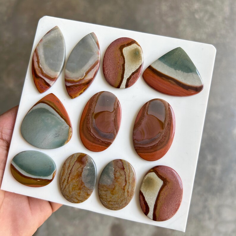 May include: Assortment of polished jasper stones in various shapes, including oval, teardrop, and triangular. The stones display a range of colors, such as red, brown, gray, and green, with unique patterns and veining. The stones are displayed on a white surface.