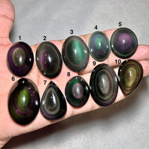 May include: Ten polished rainbow obsidian cabochons labeled one through ten, featuring concentric bands of iridescent green, purple, and gold colors. The stones are oval and teardrop-shaped with smooth, reflective surfaces.