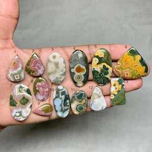 Könnte beinhalten: Eine Hand zeigt dreizehn polierte Edelsteine in verschiedenen Formen. Die Steine weisen eine Reihe von Farben auf, darunter Grün, Gelb, Braun, Rosa und Weiß, mit einzigartigen Mustern und Texturen. Die Edelsteine sind oval, tropfenförmig und dreieckig.