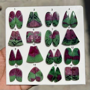 Peut inclure: Une collection de seize paires de pierres précieuses de rubis zoisite polies, de formes variées, dont des ovales, des triangles et des gouttes. Les pierres présentent un mélange de teintes rouge foncé et verte, avec des motifs et des textures visibles. Les pierres sont exposées sur une surface blanche.