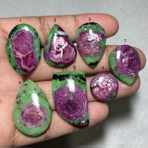 May include: Seven polished ruby zoisite gemstones in various shapes, including oval, teardrop, and round. The stones feature a vibrant mix of pink, purple, and green colors, with a polished, reflective surface.