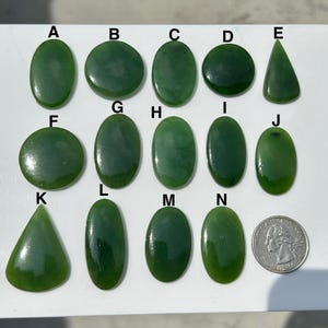 Könnte beinhalten: Eine Sammlung polierter, grüner Jadesteine in verschiedenen Formen, darunter oval, rund und dreieckig. Eine US-Viertel-Dollar-Münze ist zum Größenvergleich enthalten. Die Steine sind von A bis N nummeriert.