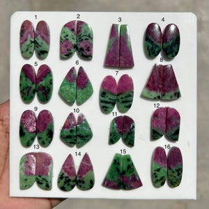 Könnte beinhalten: Eine Sammlung von sechzehn Paaren polierter Rubin-in-Zoisit-Edelsteinen, jede mit einer einzigartigen Form und Musterung. Die Steine weisen eine Mischung aus tiefen Grüntönen, leuchtendem Rosa und Schwarz auf. Die Edelsteine sind auf einer weißen Oberfläche angeordnet.