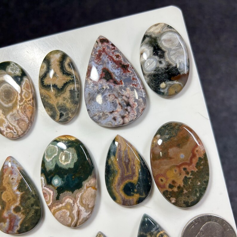 May include: A collection of polished, multi-colored ocean jasper cabochons in various oval and teardrop shapes. The stones feature intricate patterns of green, brown, cream, and red with circular orbicular inclusions, displayed on a white surface.