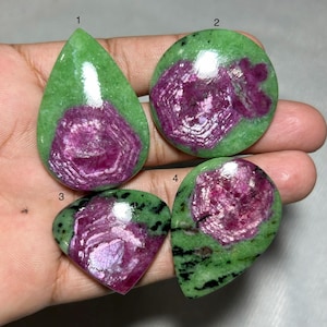 Peut inclure: Quatre pierres précieuses polies avec une matrice verte et une formation cristalline centrale magenta vif. Les pierres varient en forme : goutte d'eau, ronde et deux de forme plus angulaire. Les pierres sont tenues dans une main.