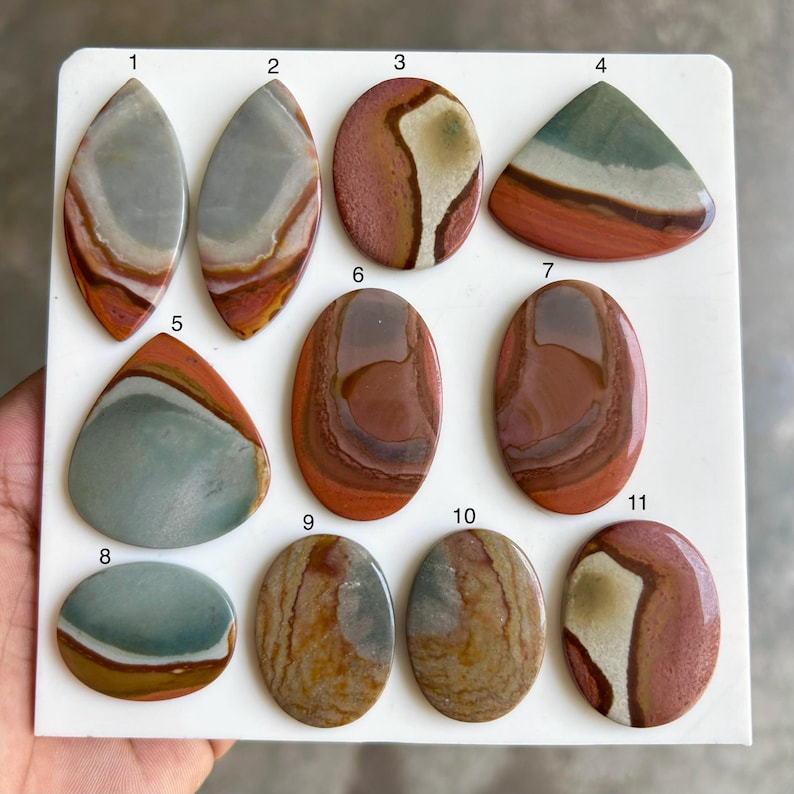May include: A collection of eleven polished gemstones in various shapes, including oval, teardrop, and triangular. The stones display a range of colors, such as red, brown, gray, and green, with unique patterns and veining. The gemstones are arranged on a white surface.