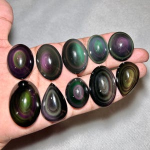 May include: A collection of ten polished rainbow obsidian cabochons in various oval and teardrop shapes. Each stone features concentric bands of iridescent green and purple hues against a dark black base, displaying a smooth, reflective surface.