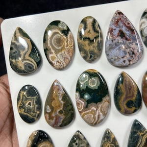 May include: A collection of polished, multi-colored cabochon gemstones in various shapes, including ovals and teardrops. The stones feature intricate, swirling patterns in shades of green, brown, cream, and grey with a smooth, reflective finish.