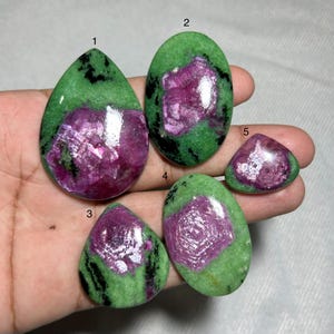 May include: Five polished gemstones in shades of green and magenta. The gemstones are polished and come in various shapes, including oval, teardrop, and triangular. The magenta centres are surrounded by green with black inclusions.