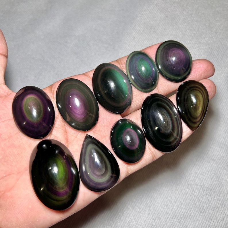 May include: A collection of ten polished rainbow obsidian cabochons in various oval and teardrop shapes. Each stone features concentric bands of iridescent green and purple hues against a deep black background with a smooth, reflective surface.