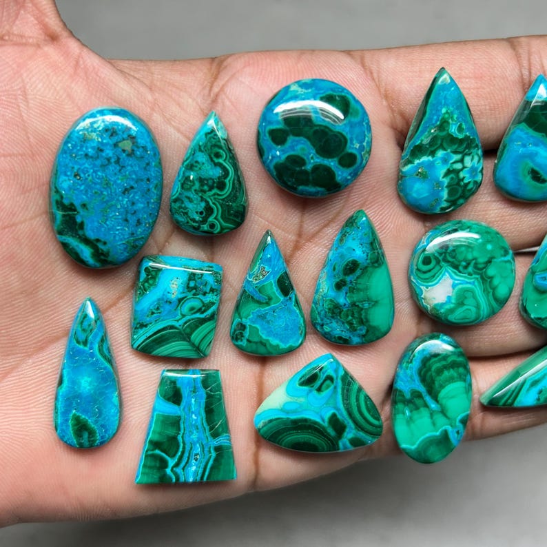 May include: A collection of polished gemstones in various shapes, including oval, teardrop, and round. The stones display a vibrant mix of turquoise and deep green hues, with intricate patterns. The gemstones are held in a hand, showcasing their natural beauty.
