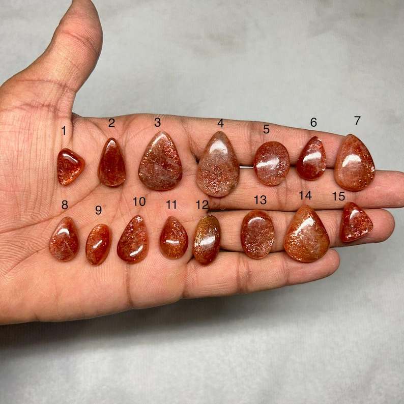 May include: A collection of fifteen polished sunstone gemstones in various shapes, including teardrop, oval, and triangular forms. The stones display a reddish-orange color with shimmering inclusions. The gemstones are held in a hand.