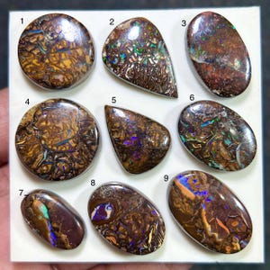 Puede incluir: Nueve ópalos boulder pulidos dispuestos en una cuadrícula, numerados del uno al nueve. Cada piedra preciosa presenta una matriz de piedra de hierro marrón oscuro con destellos iridiscentes de azul, verde y morado en sus superficies lisas.