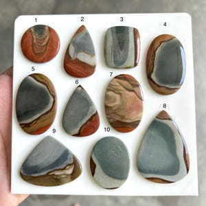 May include: A collection of polished jasper gemstones in diverse shapes, including oval, teardrop, and rectangular. The stones exhibit a blend of colours, such as grey, brown, red, and beige, with distinctive patterns and veining. Each stone is numbered.
