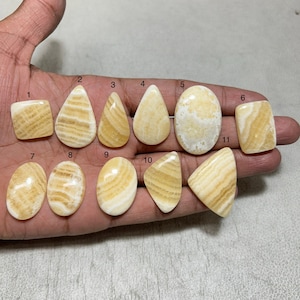 May include: A collection of eleven polished gemstones in various shapes, including squares, teardrops, and ovals. The stones display a creamy yellow color with distinctive banded patterns in shades of light brown and cream. The gemstones are displayed on a hand.