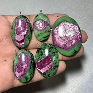 Könnte beinhalten: Fünf ovale Edelsteine mit grüner Basis und schwarzen Sprenkeln. Jeder Stein hat eine lebendige, geometrische, magenta-farbene Kristallformation in der Mitte. Die Steine werden auf hellem Hintergrund präsentiert.