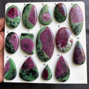 Könnte beinhalten: Eine Sammlung von vierzehn polierten Rubin-Zoisit-Cabochons, angeordnet auf einer weißen Platte und nummeriert von eins bis vierzehn. Jeder Stein zeigt eine einzigartige Kombination aus leuchtend magentafarbenen Rubineinschlüssen.