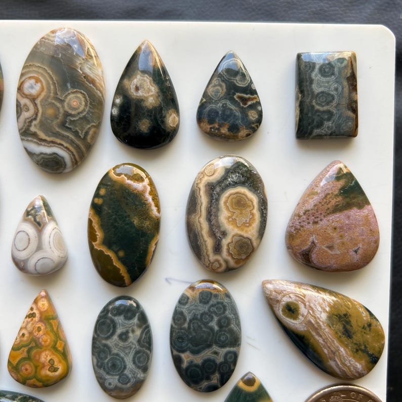 Puede incluir: Una colecci&oacute;n de cabujones de jaspe oce&aacute;nico pulidos en varias formas, incluyendo &oacute;valos, gotas y un rect&aacute;ngulo. Las piedras presentan patrones orbiculares complejos en tonos verde, marr&oacute;n, bronceado, crema y rosa con un acabado liso y brillante.