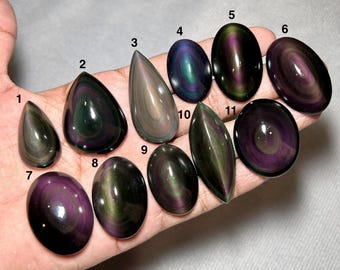 May include: Eleven polished rainbow obsidian cabochons in various shapes, including teardrop, oval, and marquise, each featuring iridescent bands of purple, green, and gold. The stones are numbered 1 through 11, displaying a smooth, reflective surface.