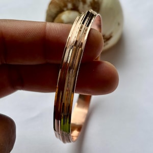 May include: A copper-colored bangle bracelet with a textured, ridged design. The bracelet has a smooth, polished finish and is held in a person's hand. The background is a blurred white.