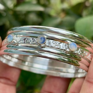 Puede incluir: Una pulsera de plata con tres pequeñas piedras preciosas redondas de color azul claro. La pulsera tiene un diseño grabado y ornamentado entre las piedras preciosas. La pulsera está hecha de múltiples bandas de plata.