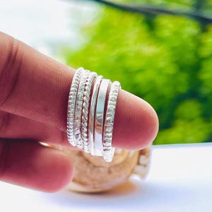 Può includere: Una collezione di anelli d'argento con varie trame, tra cui perline, intrecciati e lisci. Gli anelli sono impilati su un dito, mostrando i loro diversi stili e finiture. Lo sfondo è un verde sfocato.