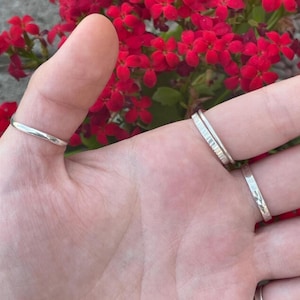 Puede incluir: Una mano con cuatro anillos de plata. Los anillos tienen diferentes diseños, incluyendo una banda lisa, una banda texturizada y una banda martillada. Flores rojas en el fondo.