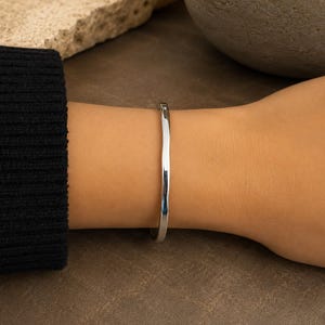 May include: A polished silver-tone metal cuff bracelet with a smooth, slightly irregular, organic wave-like texture. The bracelet features a minimalist, open-ended design that wraps around the wrist, showcasing a reflective, high-shine finish.