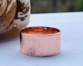 May include: A wide, copper-colored ring with a smooth, polished surface. The ring has a simple, band-style design. The ring is approximately 0.75 inches wide. The ring is a piece of jewelry.
