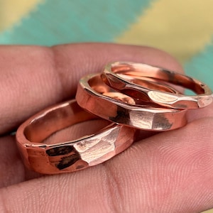 Puede incluir: Tres anillos de cobre con un acabado martillado y texturizado. Cada anillo presenta una superficie metálica pulida con facetas irregulares creadas por el proceso de martillado, mostrando un tono marrón rojizo cálido y un diseño circular.