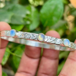 Puede incluir: Una pulsera de plata con tres cabujones redondos de piedra lunar iridiscente. La pulsera tiene un patrón floral grabado. Las piedras lunares tienen un color azul y amarillo. La pulsera está sujeta en una mano.