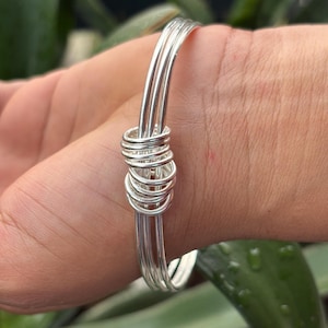 Puede incluir: Una pulsera de brazalete plateada con un diseño único. La pulsera presenta múltiples alambres paralelos unidos por una sección enrollada. La pulsera se usa en una muñeca, mostrando su estilo simple pero elegante.