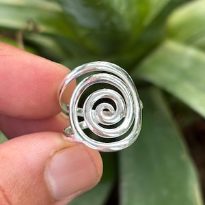 May include: A silver ring with a spiral design. The ring features a coiled, concentric pattern, creating a swirling effect. The metal has a polished, reflective surface, and the ring is held by a person's fingers.