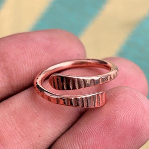 Könnte beinhalten: Ein kupferfarbener Wickelring mit einer texturierten Oberfläche, die durchgehende vertikale Einkerbungen entlang des gesamten Rings aufweist. Der Ring hat ein offenes Design mit zwei überlappenden Enden und ein metallisches Finish.
