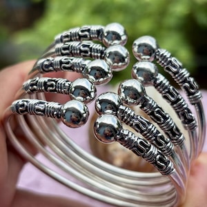 Puede incluir: Un conjunto de brazaletes de color plateado. Cada brazalete presenta un extremo liso y esférico y una sección decorativa con dibujos. Los brazaletes están apilados, creando un efecto de capas. El fondo está borroso, con toques de verde y rosa.