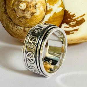Puede incluir: Anillo plateado con una banda giratoria, adornado con símbolos de la paz. El anillo tiene un acabado pulido. El fondo es una imagen borrosa de una concha marina.